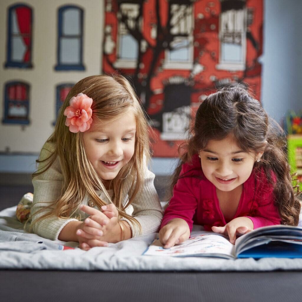 Happy Friends Reading Book While Lying On Blanket At Child Care